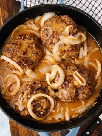 A skillet filled with four hamburger steaks topped with sautéed onions and covered in brown gravy, next to a black and white checkered towel and a knife on a wooden surface.