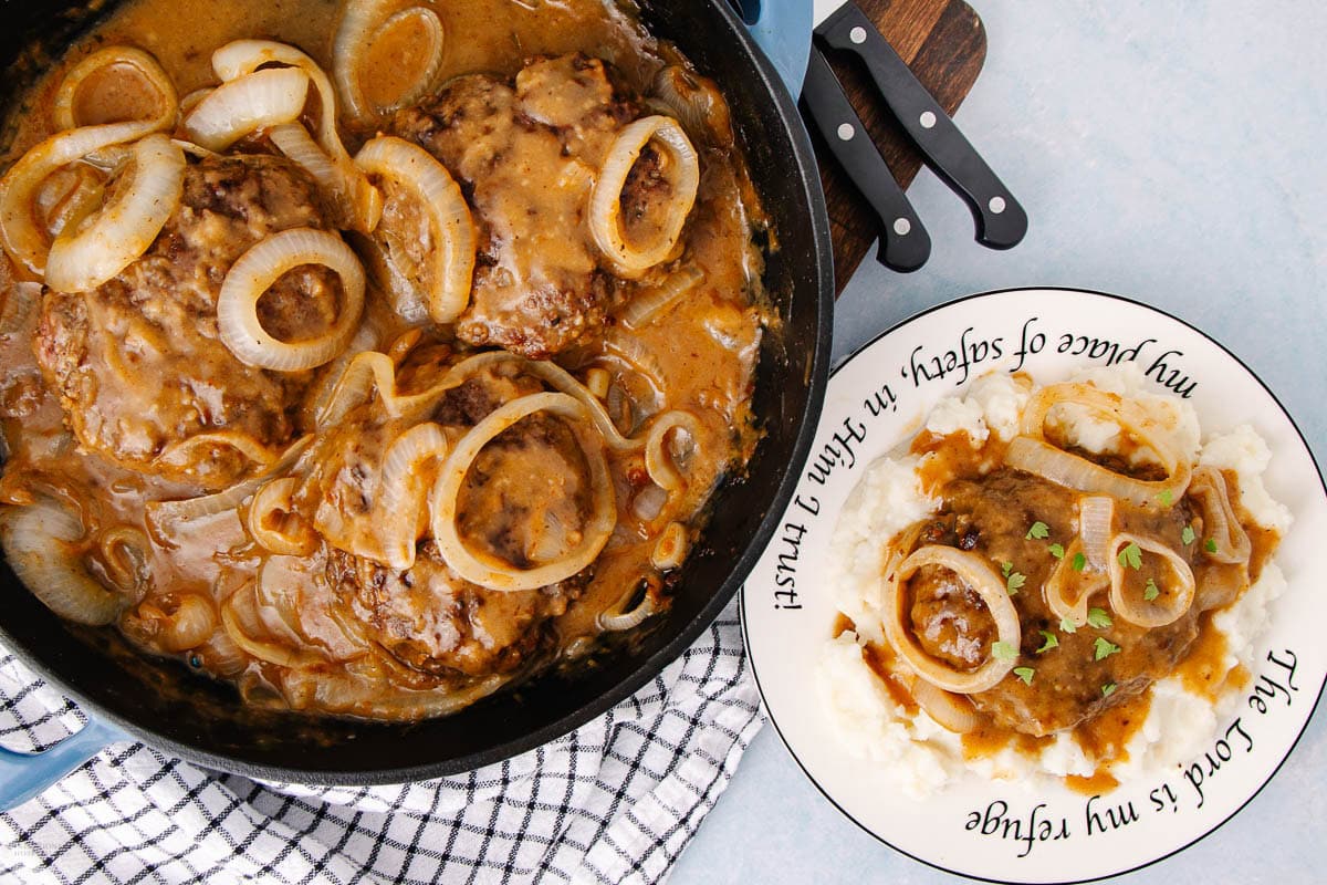 A skillet of Salisbury steak with onions and gravy next to a plate with mashed potatoes, topped with steak and onions. The plate has the phrase, "The Lord is my refuge, my place of safety, in Him I trust," written around the rim.