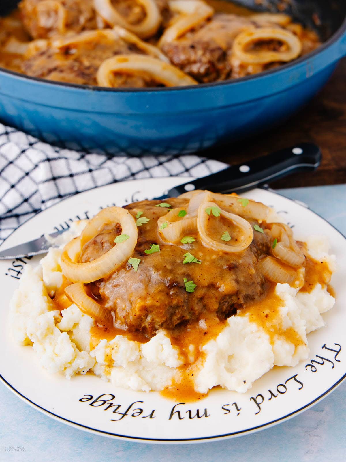 A plate of mashed potatoes topped with Salisbury steak, brown gravy, sautéed onions, and parsley sits next to a blue skillet. A black-handled fork rests on the plate, and a checkered napkin is in the background.