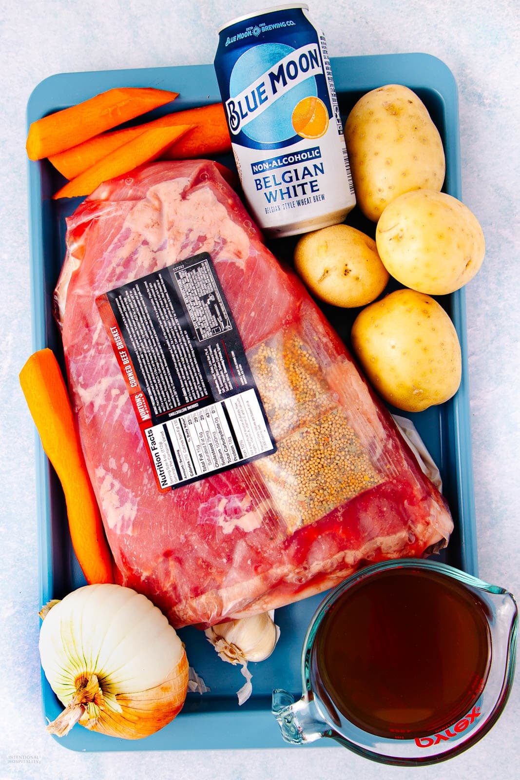 A tray holds a large raw corned beef brisket, several whole potatoes, carrots, an onion, a can of Blue Moon non-alcoholic Belgian White beer, and a measuring cup of brown liquid, likely broth.