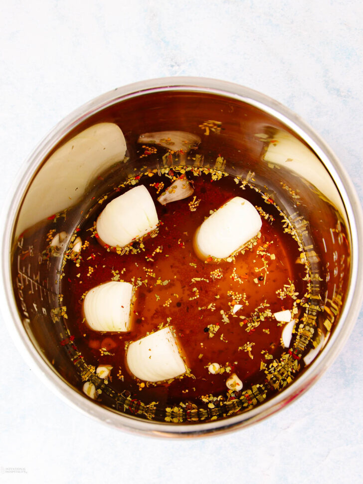 A metal pot filled with a brown liquid marinade, chunks of white onion, minced garlic, and various dried herbs and spices on a light surface.