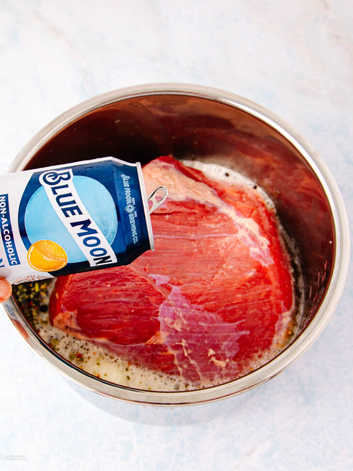 A hand pours a can of Blue Moon non-alcoholic beer into a metal bowl containing a large raw cut of beef, partially submerged in foamy liquid.