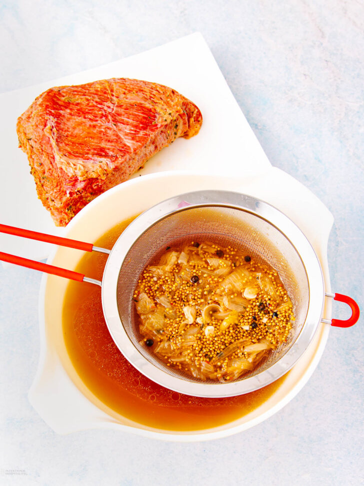 A piece of cooked corned beef sits on a white cutting board next to a bowl, where broth is being strained through a metal sieve containing mustard seeds, spices, and onions.
