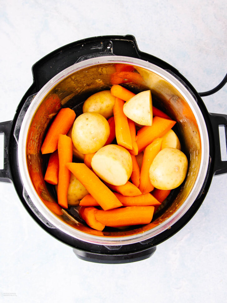 Overhead view of an electric pressure cooker filled with whole small potatoes and large carrot pieces, ready to be cooked. The vegetables are raw and brightly colored against the metal interior.