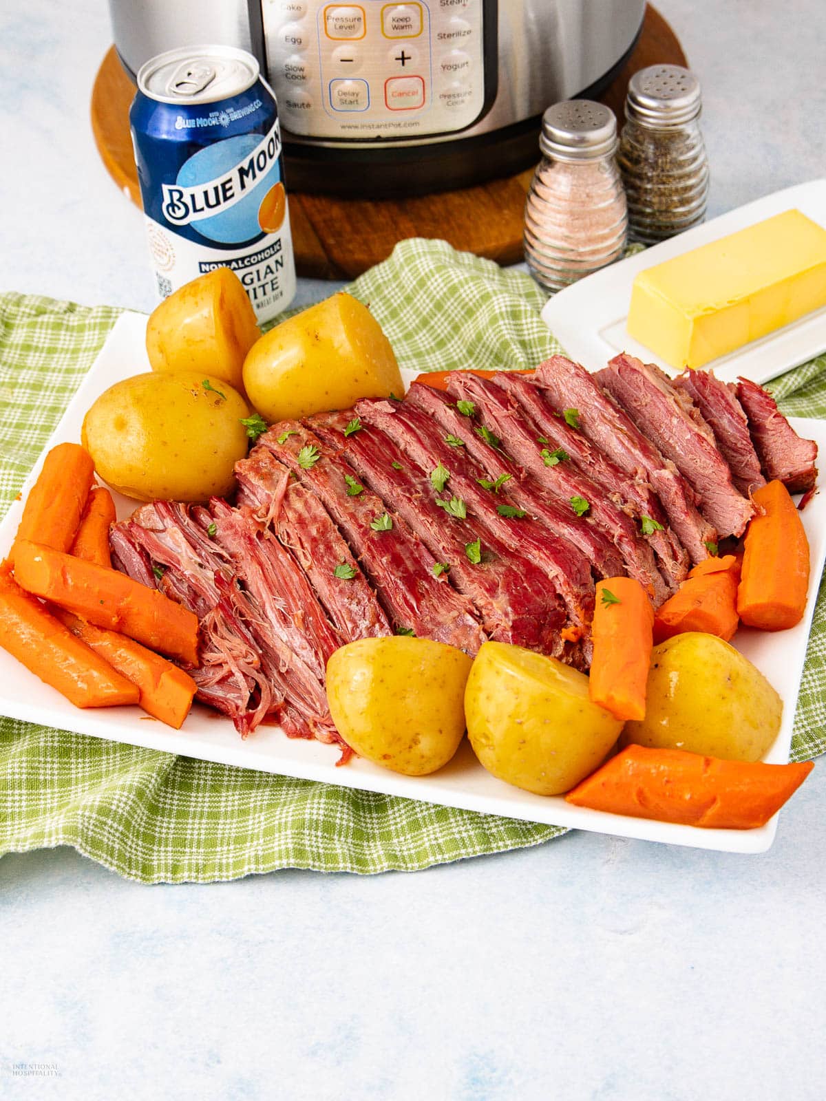 A platter of sliced corned beef garnished with parsley, surrounded by cooked carrots and yellow potatoes. Nearby are a stick of butter, a salt and pepper shaker, a can of Blue Moon beer, and an Instant Pot in the background.