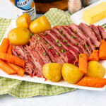 Sliced corned beef brisket garnished with herbs, served with whole yellow potatoes and carrot pieces on a white platter, set on a green checkered cloth with butter, salt, and pepper in the background.