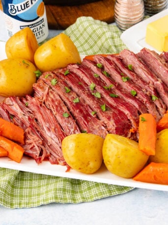 Sliced corned beef brisket garnished with herbs, served with whole yellow potatoes and carrot pieces on a white platter, set on a green checkered cloth with butter, salt, and pepper in the background.