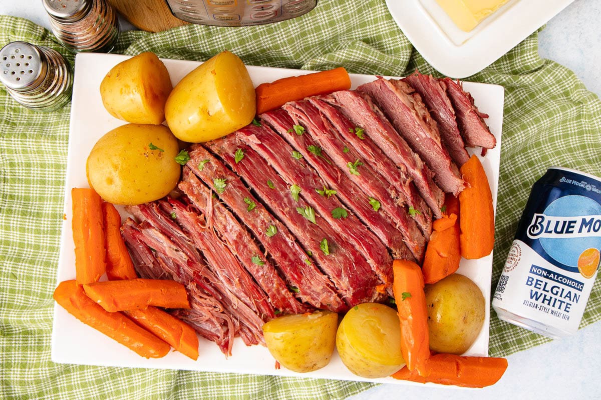 A platter of sliced corned beef garnished with parsley, surrounded by whole yellow potatoes and carrots. A can of Blue Moon Belgian White beer sits nearby on a green checkered tablecloth.