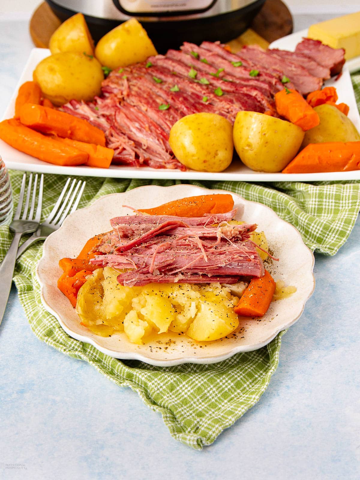 A plate of corned beef, potatoes, and carrots sits on a green checkered napkin, with a larger platter of the same meal and an Instant Pot in the background. The food is garnished with herbs.