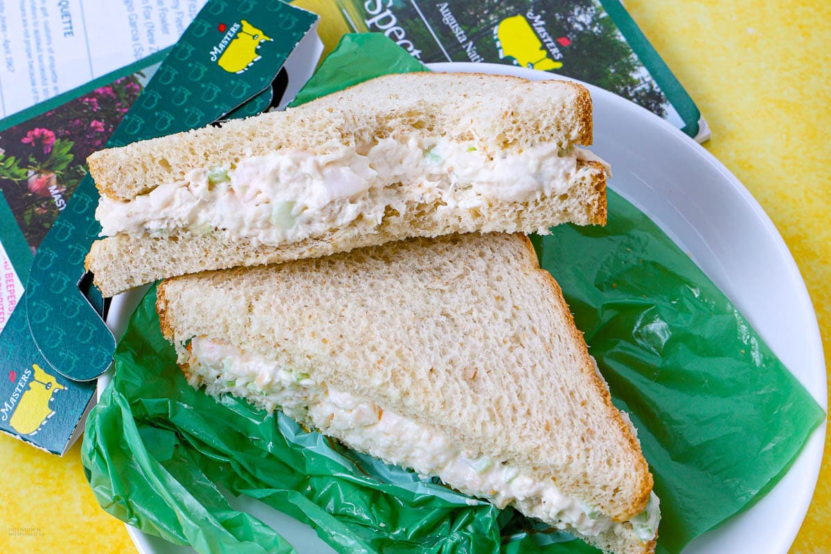 A plate with a chicken salad sandwich cut in half on white bread, placed on green wax paper, with travel brochures partially visible in the background.