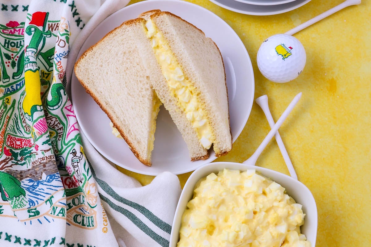 A plate with a halved egg salad sandwich on white bread sits next to a bowl of egg salad, a golf ball, tees, and a colorful towel on a yellow surface.