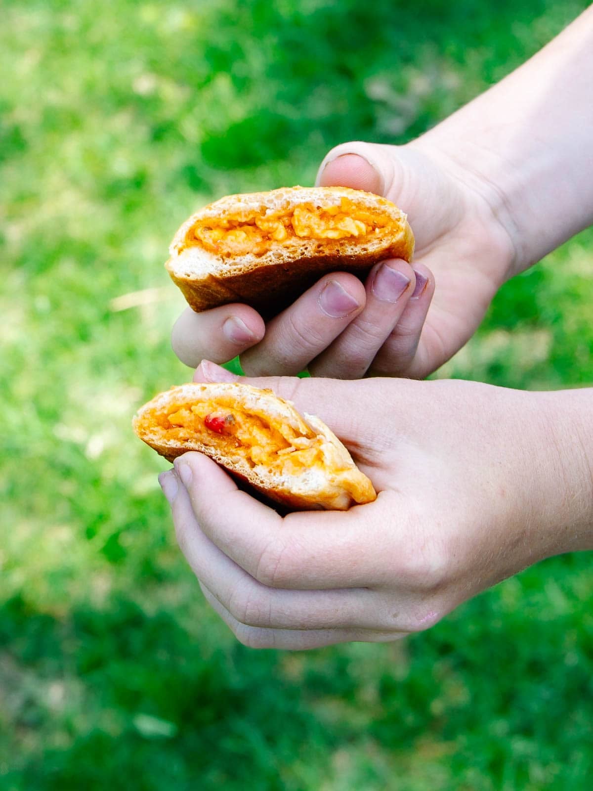 Two hands holding halves of a bread roll filled with melted orange cheese, with green grass in the background.