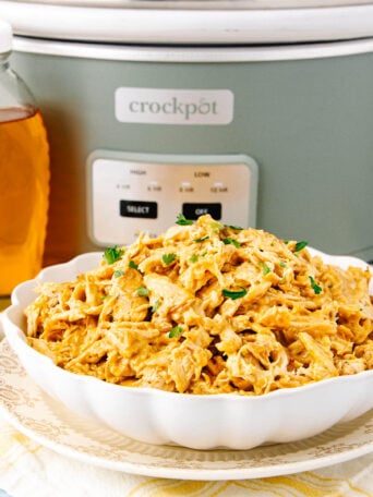 A white bowl filled with creamy shredded chicken sits on a plate in front of a green Crockpot. A glass jar of honey and a small dish of mustard are beside it. The chicken is garnished with chopped herbs.