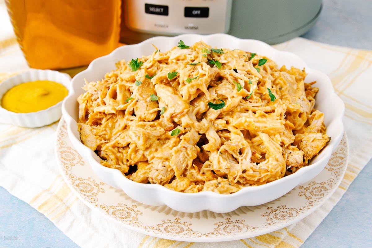 A white scalloped bowl filled with shredded chicken coated in a creamy sauce, garnished with chopped herbs, sits on a patterned plate. There is a small dish of mustard and a kitchen appliance in the background.