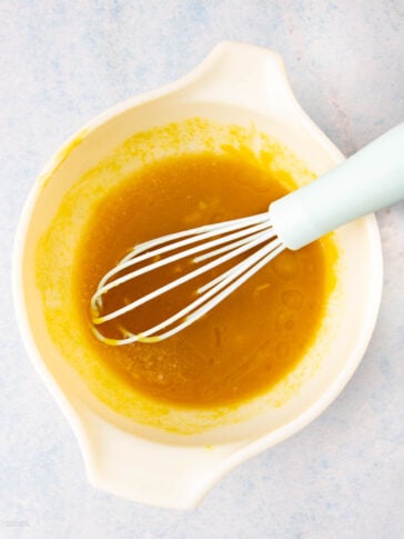 A white mixing bowl with a light blue-handled whisk stirring a yellow liquid mixture, set on a light blue surface.