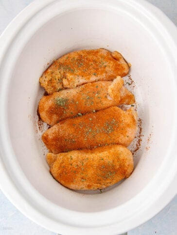 Four raw chicken breasts coated with seasoning and herbs are arranged in a white slow cooker, ready to be cooked. The seasoning appears to include paprika and dried green herbs.