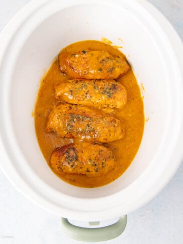 Four cooked chicken breasts sit in a white slow cooker, covered in a thick, orange-brown sauce with visible herbs and seasonings. The sauce surrounds the chicken at the bottom of the cooker.