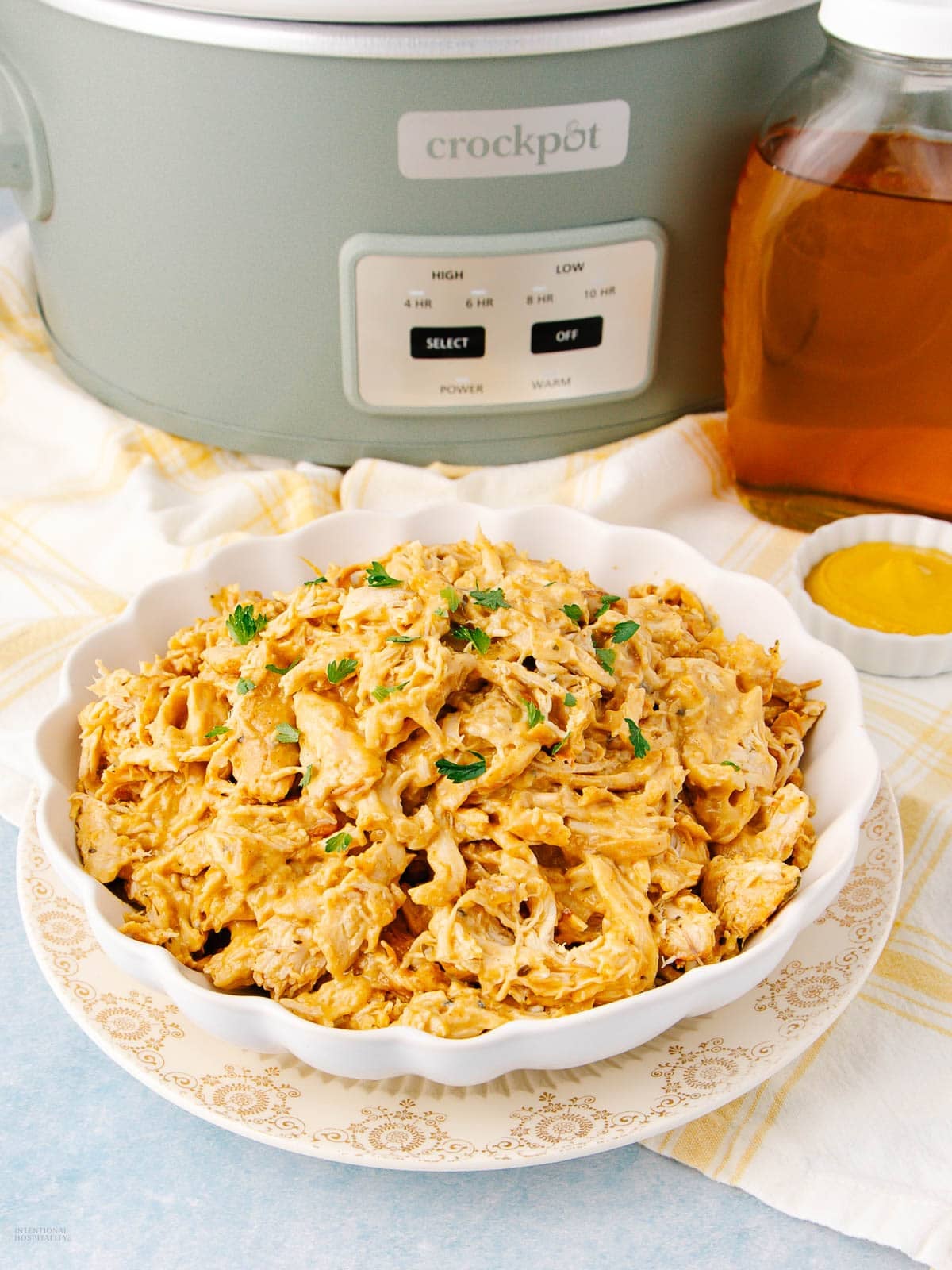 A bowl of shredded barbecue chicken garnished with herbs sits on a patterned plate in front of a crockpot, with a jar of sauce and a small dish of mustard nearby on a yellow-checked cloth.