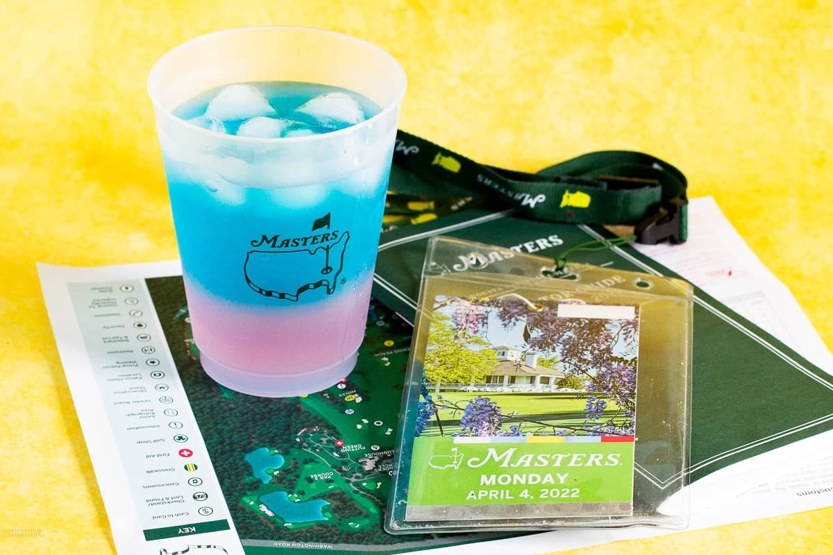 A plastic Masters cup with a blue and pink drink and ice sits on a Masters event map next to a 2022 Masters Monday ticket badge and a green lanyard, all on a yellow surface.