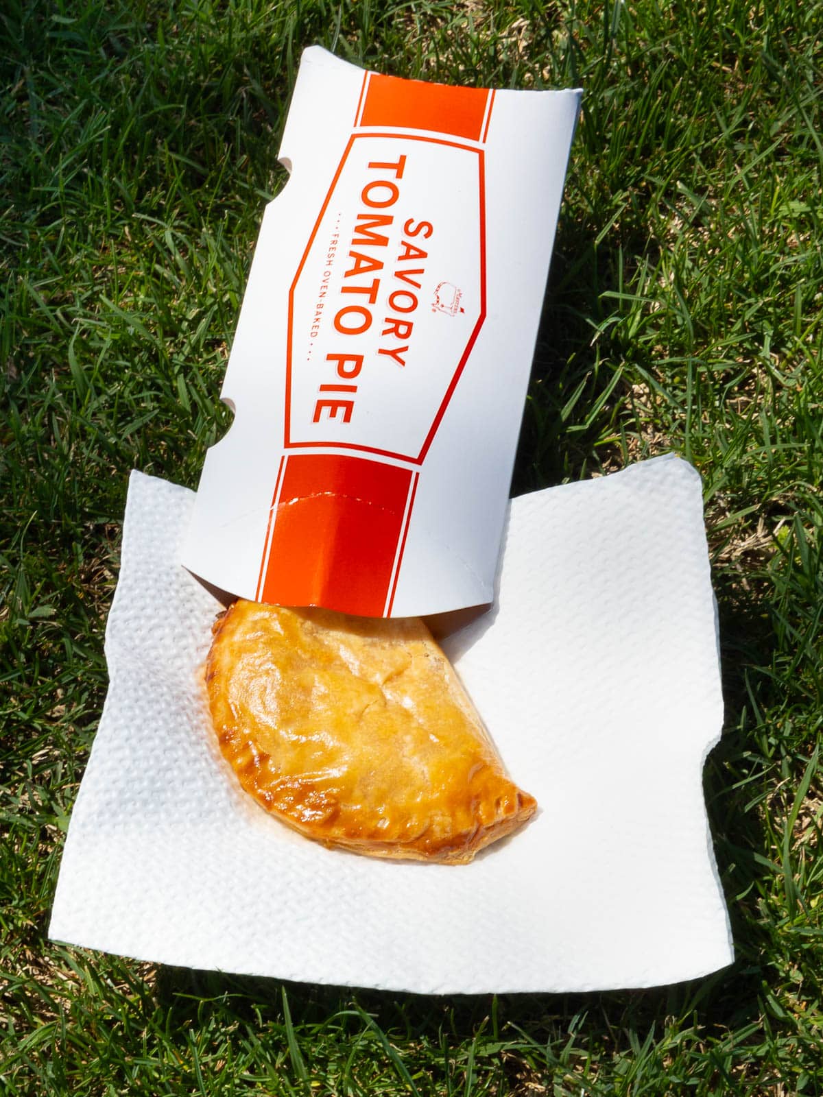 A golden-brown savory tomato pie in a red and white paper sleeve rests on a white napkin on grass, with sunlight highlighting its flaky crust.