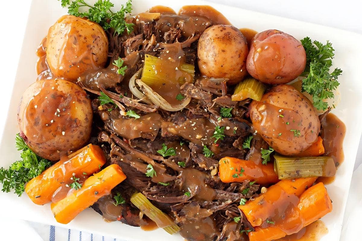 A platter of pot roast with shredded beef, whole potatoes, carrots, and celery, all topped with brown gravy and garnished with fresh parsley.