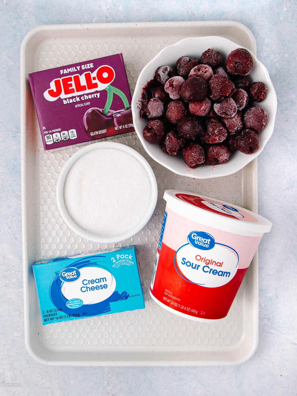A tray with a box of black cherry Jello, a bowl of frozen dark cherries, a bowl of sugar, a package of cream cheese, and a container of sour cream arranged on a light surface.