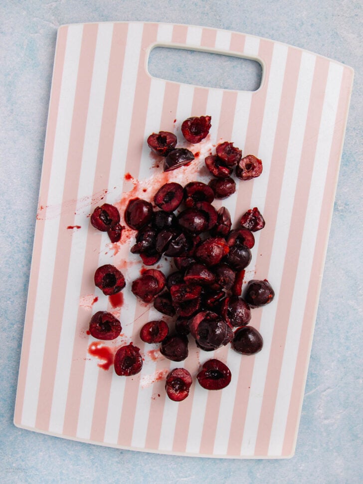 A white and light pink striped cutting board with halved and pitted dark red cherries scattered across it, some juice staining the board.