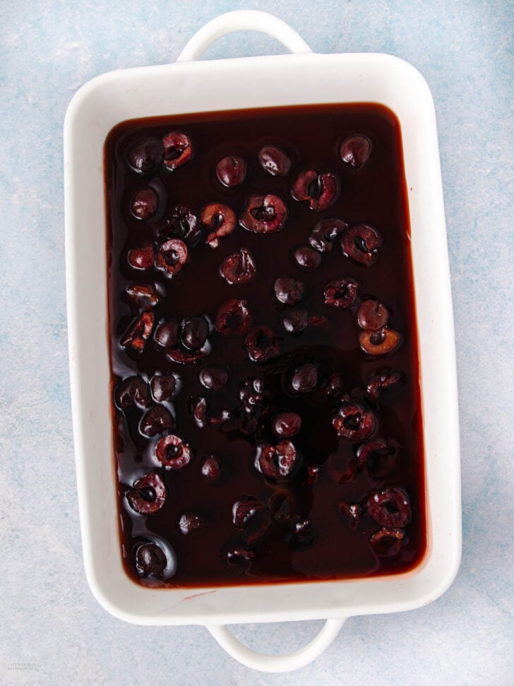 A white rectangular baking dish filled with dark cherries in a rich, glossy sauce, placed on a light blue textured surface.