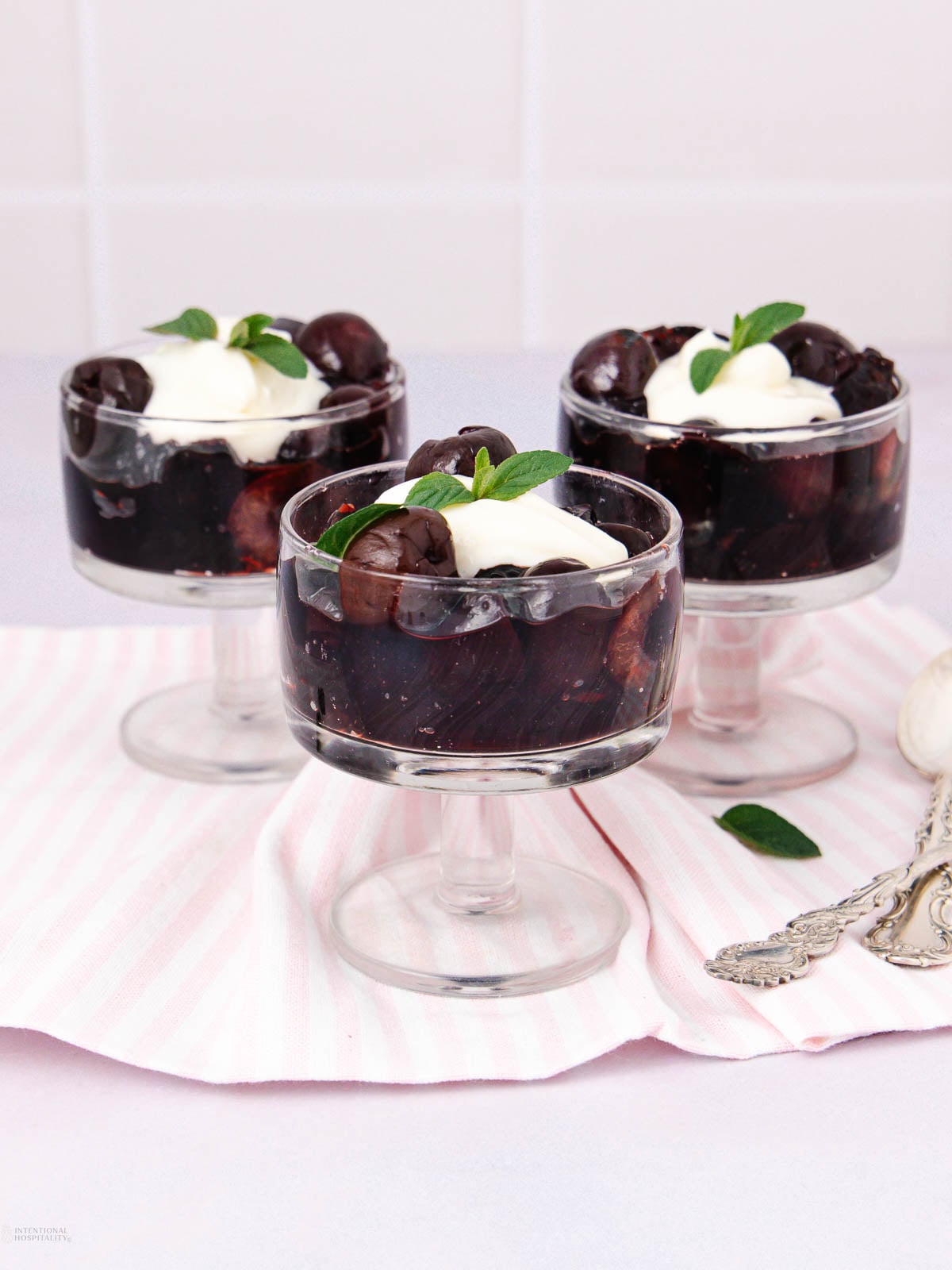 Three glass dessert cups filled with dark cherries, topped with whipped cream and mint leaves, are arranged on a pink-striped cloth with two ornate silver spoons beside them.