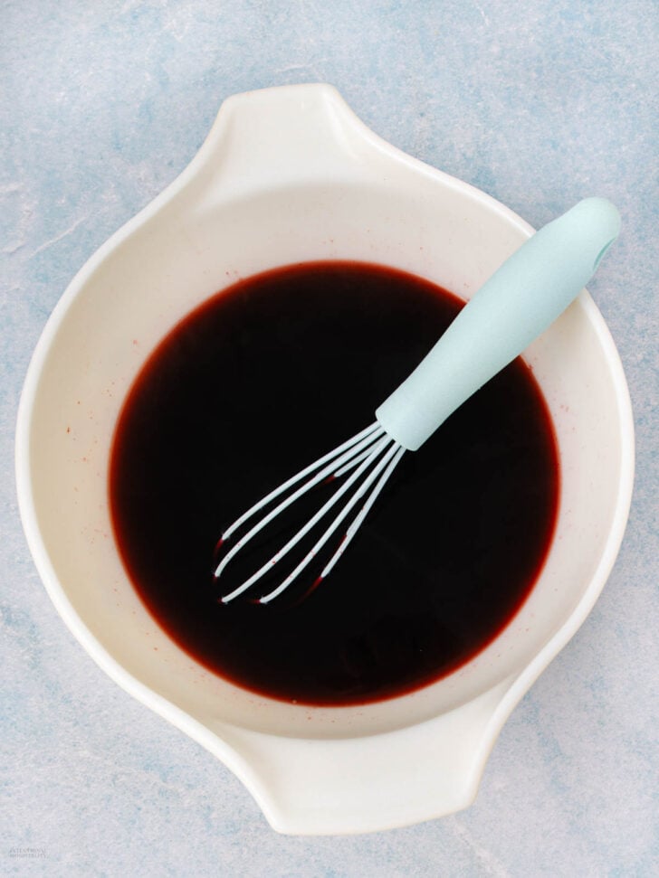 A white mixing bowl filled with a dark red liquid, with a light blue-handled whisk resting inside, placed on a light blue textured surface.