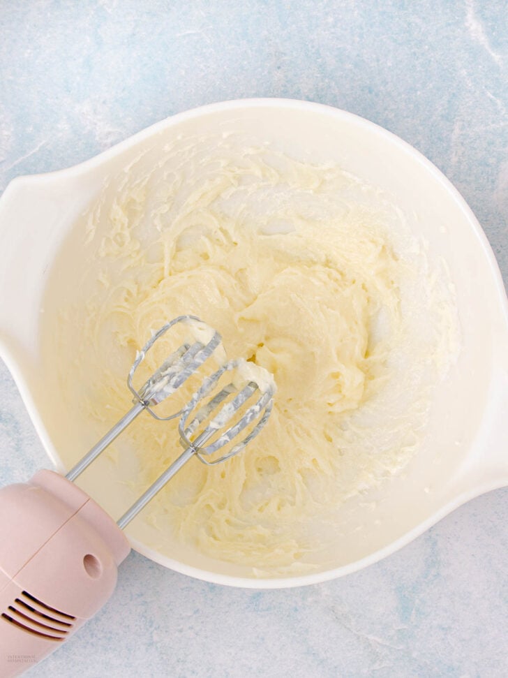 A white mixing bowl filled with creamy, whipped mixture, with a pink electric hand mixer resting inside, on a light blue surface.