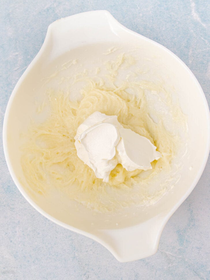 A white mixing bowl containing a creamy, pale yellow mixture with two dollops of white cream on top, set on a light blue surface.