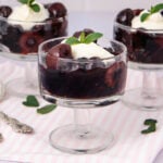 Three glass dessert cups filled with dark cherry compote, topped with a dollop of whipped cream and a mint leaf, placed on a light pink striped cloth with scattered mint leaves nearby.