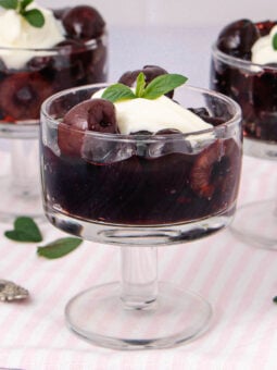 Three glass dessert cups filled with dark cherry compote, topped with a dollop of whipped cream and a mint leaf, placed on a light pink striped cloth with scattered mint leaves nearby.