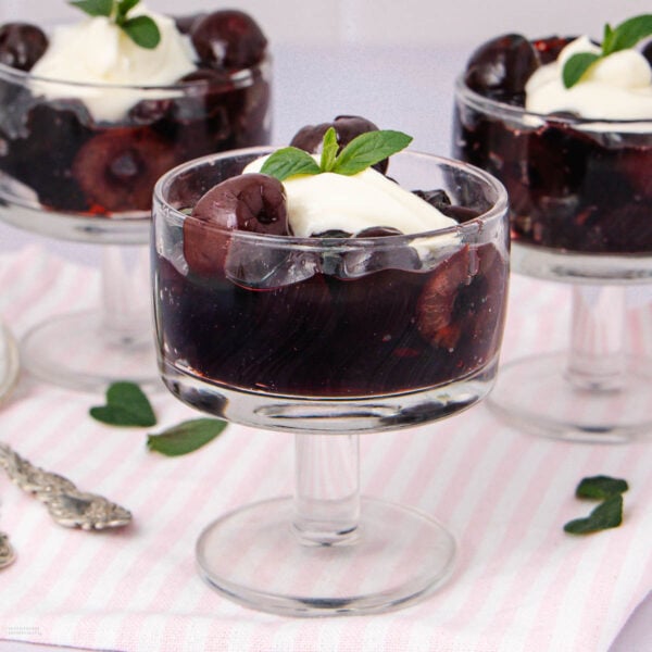 Three glass dessert cups filled with dark cherry compote, topped with a dollop of whipped cream and a mint leaf, placed on a light pink striped cloth with scattered mint leaves nearby.