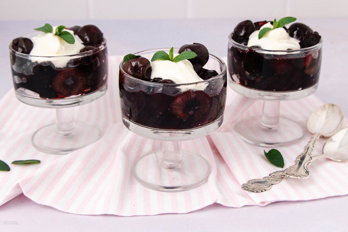 Three glass dessert cups filled with dark cherries and topped with whipped cream and mint leaves, displayed on a pink and white striped cloth with vintage silver spoons nearby.