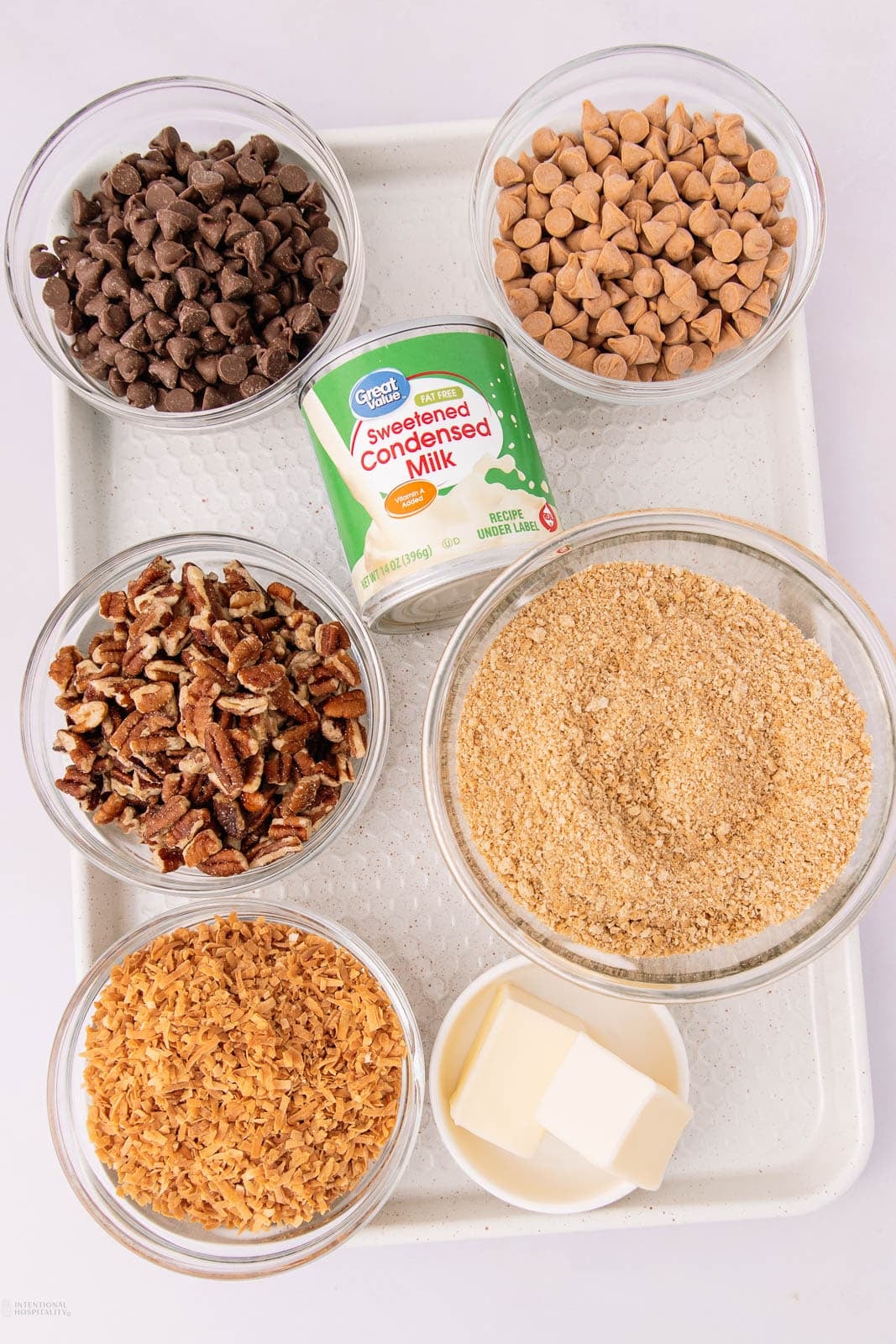 A tray with bowls containing chocolate chips, butterscotch chips, chopped pecans, graham cracker crumbs, shredded coconut, butter, and a can of sweetened condensed milk.