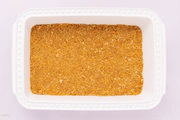 A white rectangular baking dish filled with an even layer of golden brown, crumbly crust mixture, likely made from crushed cookies or graham crackers.