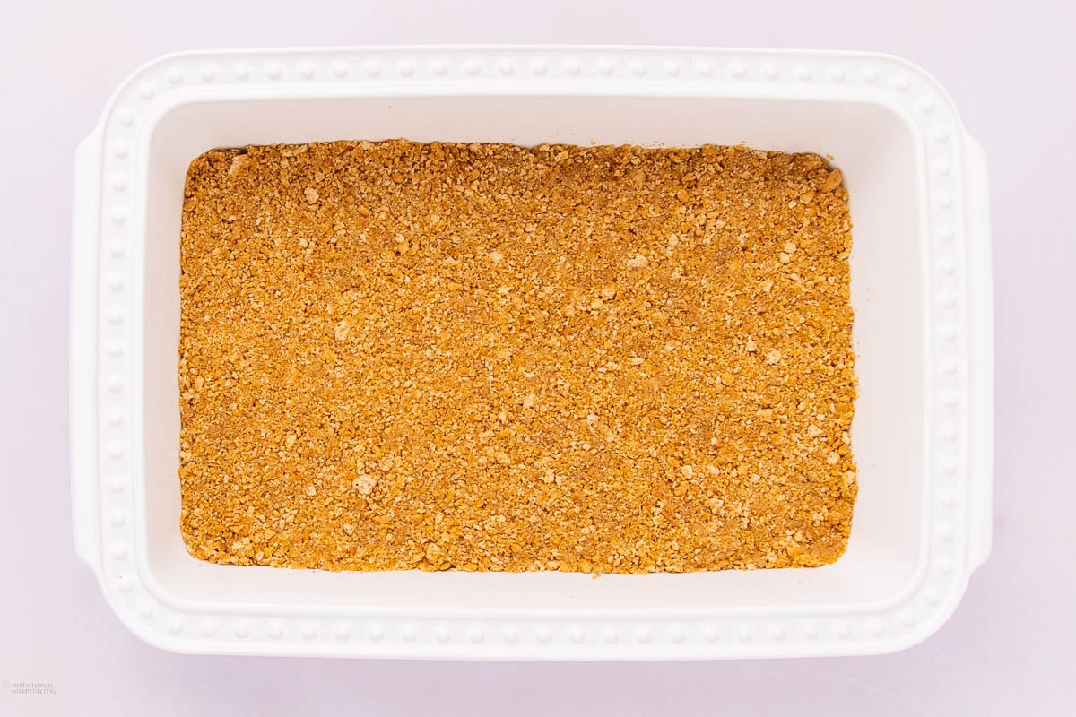 A white rectangular baking dish filled with an even layer of golden brown, crumbly crust mixture, likely made from crushed cookies or graham crackers.