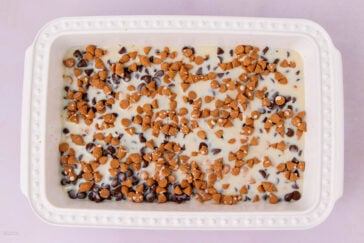 A rectangular white baking dish filled with a mixture of sweetened condensed milk, chocolate chips, and butterscotch chips, evenly distributed on top, on a light purple background.