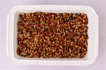 A white rectangular baking dish filled with a mixture of chopped pecans, chocolate chips, and butterscotch chips on a light pink surface.