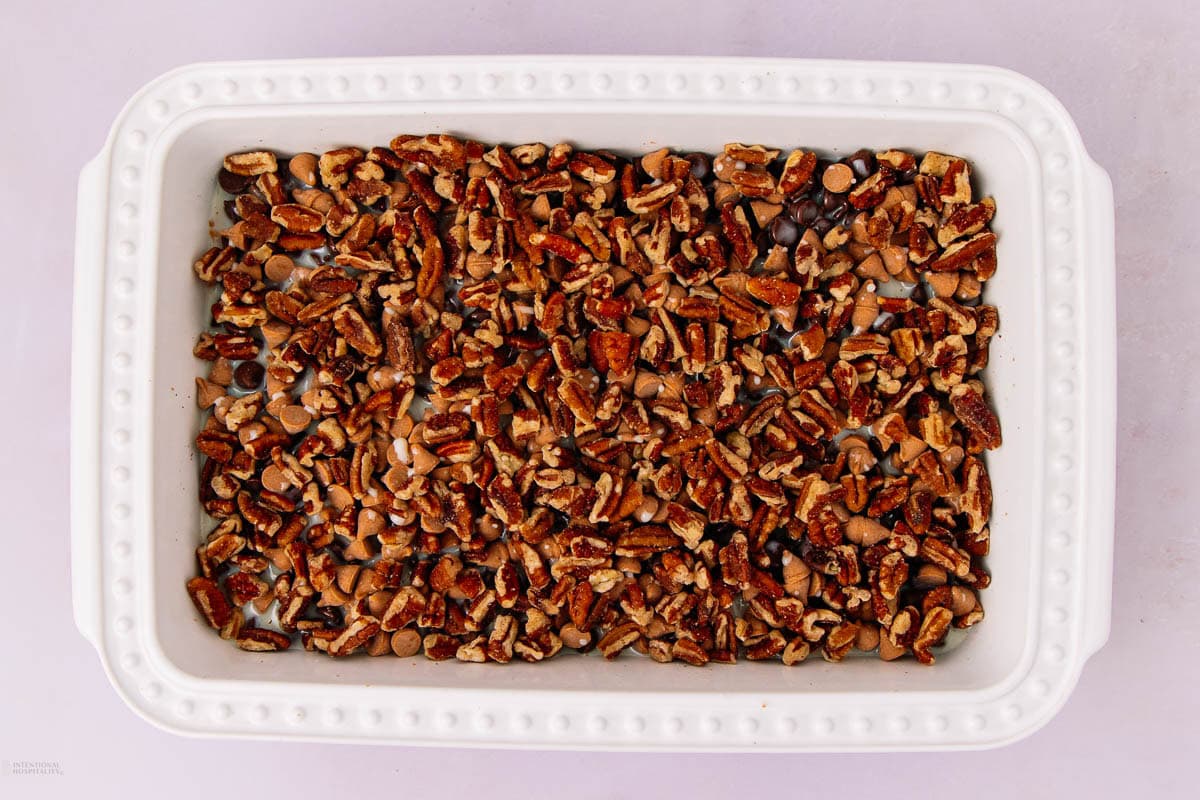 A white rectangular baking dish filled with a mixture of chopped pecans, chocolate chips, and butterscotch chips on a light pink surface.