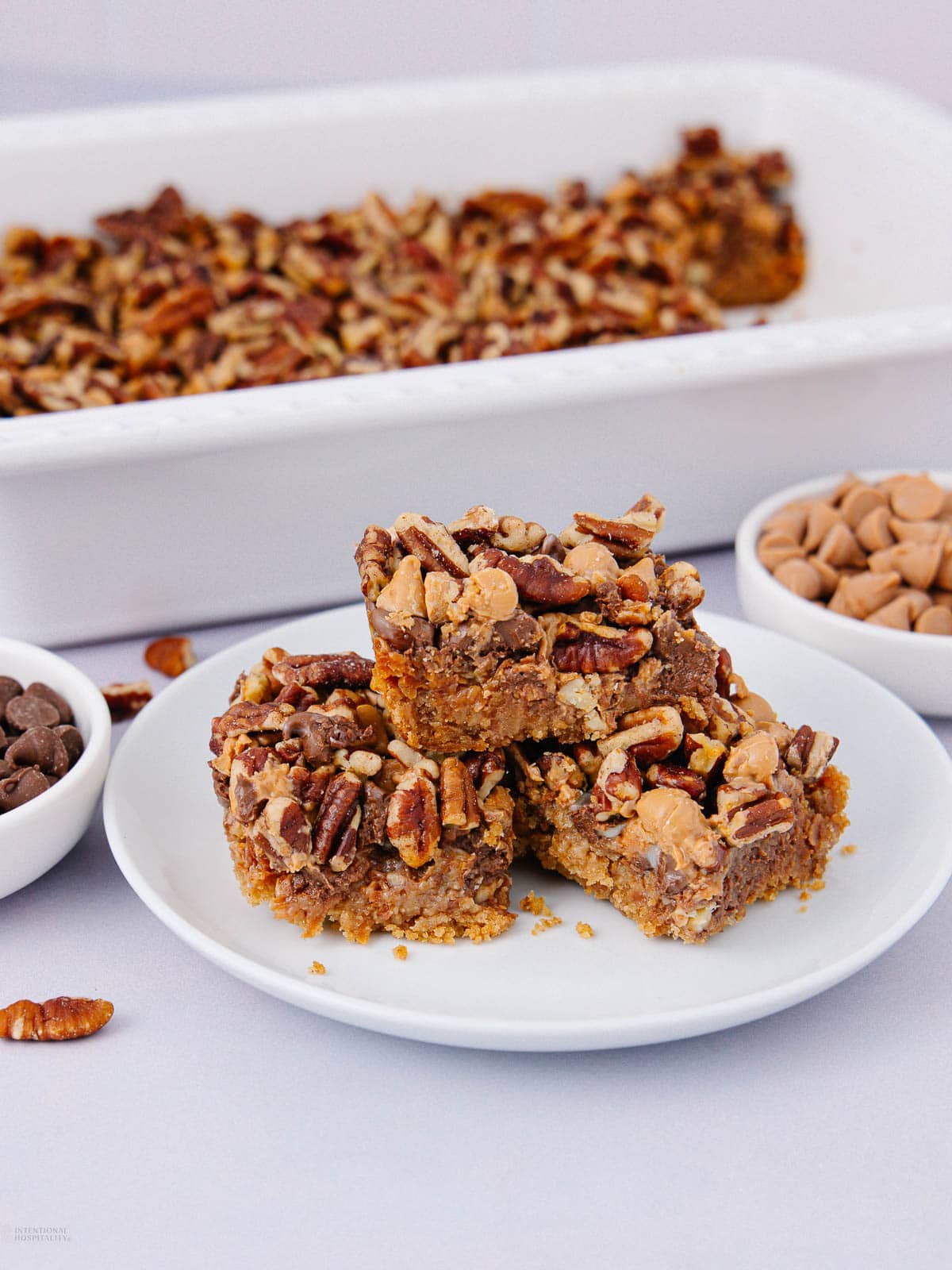 Three pecan bars stacked on a white plate, topped with chopped pecans and butterscotch chips. In the background, a baking dish with more bars and small bowls of butterscotch chips, chocolate chips, and pecans.