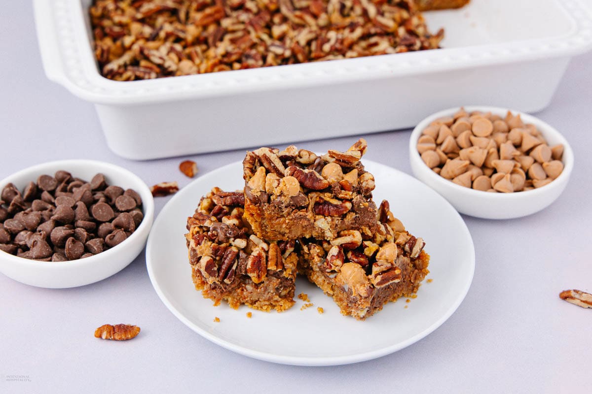 A plate with three nutty dessert bars sits in front of a baking dish of more bars. The plate is surrounded by small bowls of chocolate chips, peanut butter chips, and chocolate chips. Pecan pieces are scattered around.