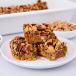 Three pecan bars stacked on a white plate, with a crumbly base and topped with chopped pecans and butterscotch chips. A dish of bars and a bowl of butterscotch chips are visible in the background.