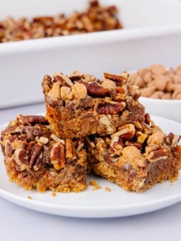 Three pecan bars stacked on a white plate, with a crumbly base and topped with chopped pecans and butterscotch chips. A dish of bars and a bowl of butterscotch chips are visible in the background.