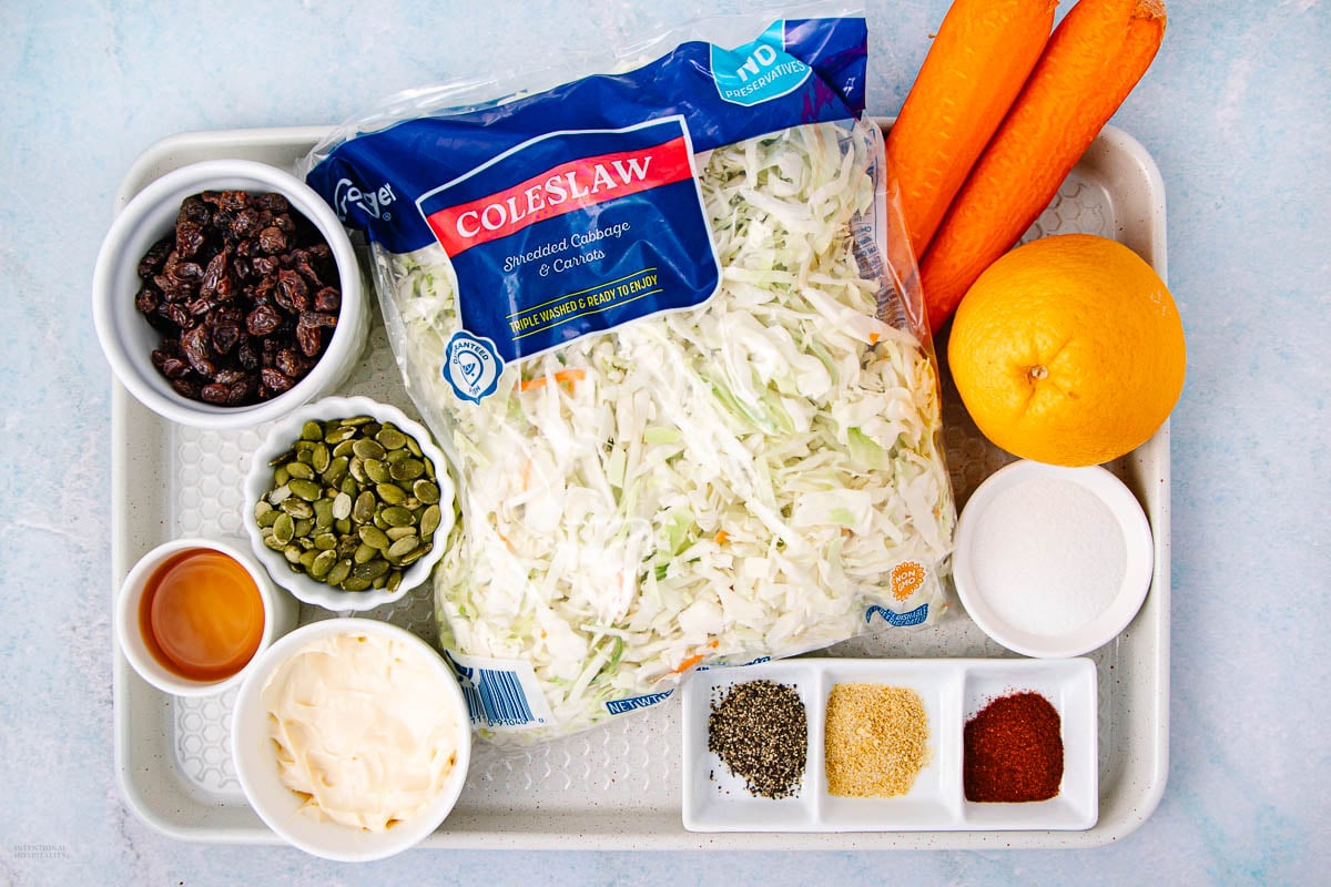 A tray with ingredients for coleslaw: bag of shredded cabbage, two carrots, an orange, small bowls of raisins, pepitas, mayonnaise, honey, sugar, ground pepper, granulated garlic, paprika, and salt.