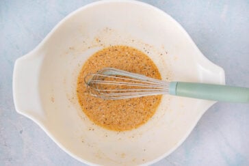 A metal whisk with a green handle rests in a large white mixing bowl containing a seasoned mixture of liquid and spices on a light blue surface.