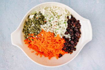 A white bowl containing shredded cabbage, grated carrots, pumpkin seeds, and raisins, all arranged in separate sections on a light blue surface.