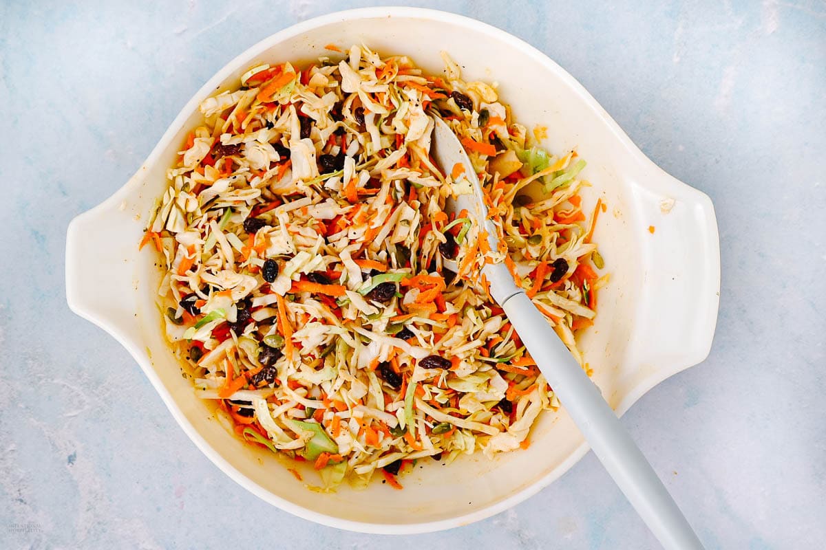 A white bowl filled with coleslaw made of shredded cabbage, carrots, raisins, and possibly other vegetables, with a serving spoon resting inside, on a light blue surface.